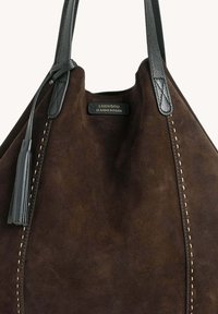 Sac en suède marron foncé avec des coutures contrastantes en crème, doté de poignées en cuir noir et d'un accent en pampille. Étiquette de logo visible en haut.