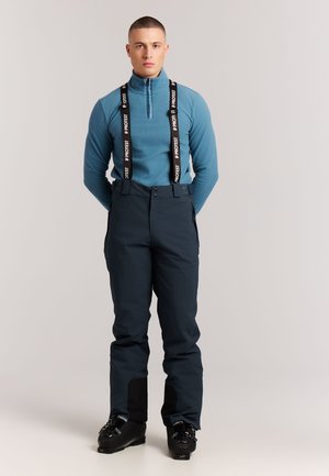 Homme en haut bleu à manches longues et pantalon de neige foncé avec des bretelles de marque et des chaussures de ski noires, debout devant un fond uni.
