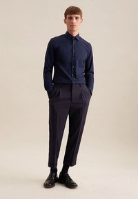 Marineblauw overhemd met knopen en een puntige kraag, gecombineerd met donkere, taps toelopende broek. Zwarte leren schoenen maken de outfit compleet. Gladde stoftextuur.