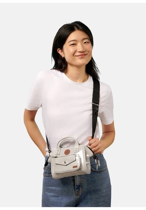 Junge Frau lächelnd, trägt ein weißes T-Shirt und Jeans, trägt eine kleine silberne Handtasche mit schwarzem Riemen über der Schulter.