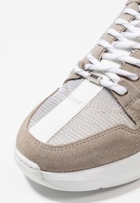 Zamszowy i tekstylny sneaker w jasnoszarej i białej kolorystyce. Wyposażony w płaskie białe sznurówki oraz metaliczne logo po boku.