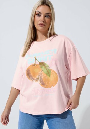 Blondynka w luźnej różowej koszulce z grafiką pomarańczy oraz jasnoniebieskich dżinsach, stojąca na tle jednolitego jasnego tła.
