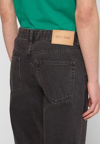 Pantalones de mezclilla negros con corte recto, que cuentan con dos bolsillos traseros y un parche de cuero marrón claro etiquetado "ONLY & SONS" en la cintura.
