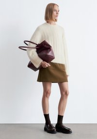 Cremefarbener Strickpullover mit strukturiertem Finish, olivgrüner Rock und dunkelbraune Loafers mit dicken Sohlen. Taktile burgunderrote Tragetasche in der Hand.