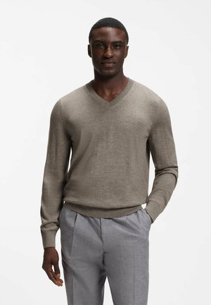 Mann trägt einen beigen V-Ausschnitt-Pullover und hellgraue Hose, steht mit einer Hand in der Tasche vor einem schlichten weißen Hintergrund.