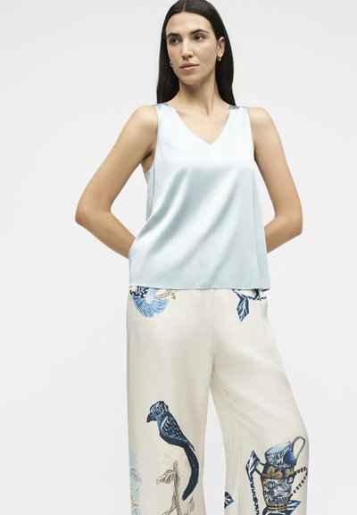 Femme aux cheveux foncés portant un haut sans manches bleu clair et un pantalon crème avec des imprimés d’oiseaux bleus et marron ainsi que des motifs floraux, debout avec les mains dans le dos.