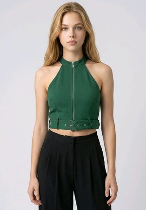 Joven mujer con pelo largo y rubio lleva un top corto verde sin mangas con cremallera frontal y cinturón, combinado con pantalones negros de cintura alta.