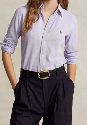 Femme portant une chemise boutonnée violet clair avec les manches retroussées, rentrée dans un pantalon noir taille haute avec une ceinture noire et une boucle dorée.