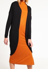 En kvinna bär en svart öppen cardigan över en ribbad, orange midiklänning och står framför en enfärgad vit bakgrund.