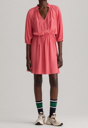 Mannequin portant une robe rose corail longueur genou avec manches 3/4, taille élastique, encolure à nouer, chaussettes rayées et baskets blanches.