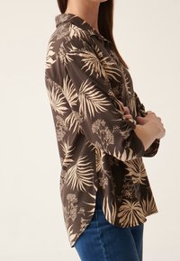 Chemise marron à manches longues ornée d'un motif botanique beige. Le tissu a une texture lisse, avec un col et des fentes sur les côtés.