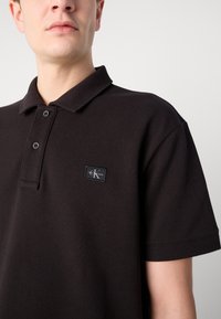 Svart polo t-skjorte laget av teksturert stoff, med klassisk krage, to-knappsspalt og en liten merketikett på brystet.