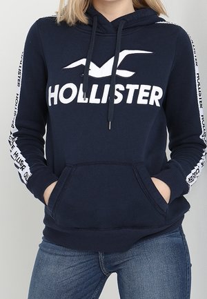 Osoba nosząca granatową bluzę Hollister z kapturem z logo nadrukowanym na piersi i rękawach, ręce w przedniej kieszeni, w połączeniu z niebieskimi dżinsami.