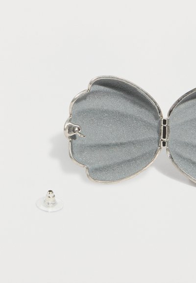 Ασημένιο πίσω μέρος σκουλαρικιού σε σχήμα κοχυλιού με αποσπώμενο διάφανο πλαστικό στοπ σκουλαρικιού σε λευκό φόντο.