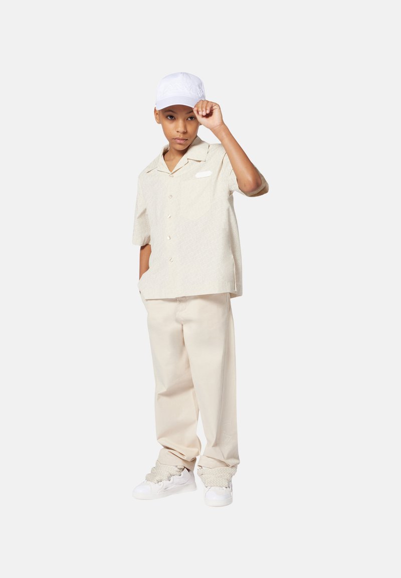 Lichtbeige overhemd met korte mouwen en een subtiel patroon, gecombineerd met bijpassende beige broek en witte sneakers. Witte pet schuin gedragen.