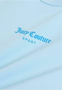 Világoskék pamutpóló, amelyen a "Juicy Couture" szöveg kék, dőlt betűkkel szerepel, alatta pedig kisebb betűkkel a "SPORT" felirat található.
