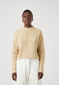 Maglione a coste beige con scollatura ampia e vestibilità rilassata. Presenta un logo "CK" in rilievo a contrasto sul davanti.
