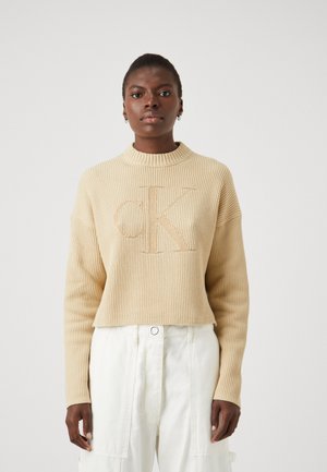 Maglione a coste beige con scollatura ampia e vestibilità rilassata. Presenta un logo "CK" in rilievo a contrasto sul davanti.