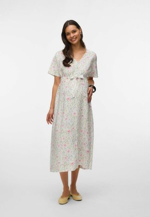 Smilende kvinde med langt mørkt hår bærer en lys blomsterprintet midi-kjole med taljebånd, beige flats og et sort armbånd, stående foran en hvid baggrund.