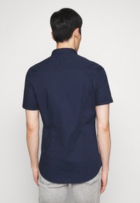 Uomo che indossa una camicia aderente a maniche corte di colore navy e jeans grigio chiaro, in piedi con la schiena rivolta verso la fotocamera su uno sfondo neutro.