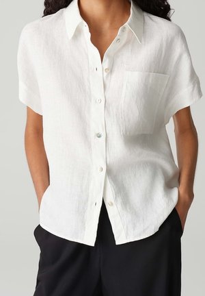 Femme portant une chemise en lin blanche à manches courtes boutonnée à l'avant avec une poche poitrine, associée à un pantalon noir, mains dans les poches.