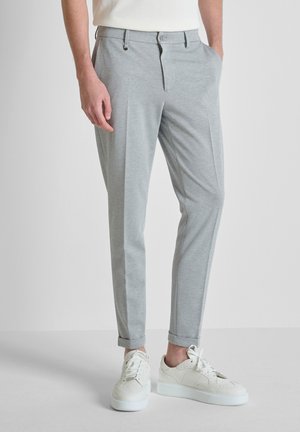 Uomo che indossa pantaloni aderenti grigio chiaro con orli risvoltati e sneakers bianche basse, in piedi con una mano in tasca.