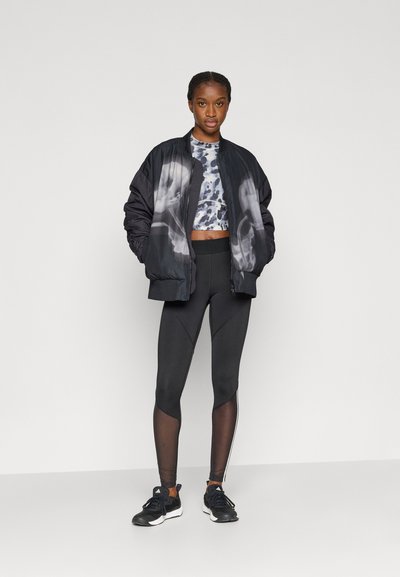 Μαύρο μπουφάν μπόμπερ με διαβαθμισμένο σχέδιο, cropped τοπ με σχέδια, μαύρο κολάν με διακοσμητικές διαφάνειες από δίχτυ και μαύρα αθλητικά παπούτσια.