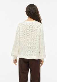 Femme aux longs cheveux bouclés portant un pull en tricot blanc avec un motif géométrique en dentelle et un pantalon marron foncé, vue de dos.