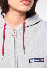 Felpa grigia con zip, dotata di corde rosse a puntini blu, un tirante per la zip e un patch con logo "ellesse" blu e rosso sul petto.