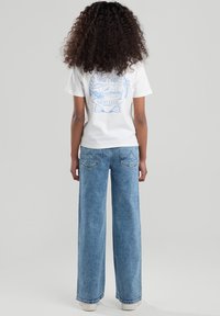 Valkoinen puuvillapaita, jossa on sininen graafinen kuvasto selkäosassa, yhdistettynä vaaleansinisiin leveälahkeisiin farkkuihin, joissa on taskut ja suora istuvuus.