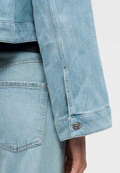Μανίκι ανοιχτού μπλε τζιν μπουφάν με λεπτομέρεια μεταλλικού κουμπιού και τσέπη από τζιν του ίδιου χρώματος εμφανής στο χέρι και το ισχίο ενός ατόμου.