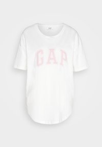 Camiseta de algodón blanca de manga corta que presenta un logo rosa de "GAP" en la parte frontal, cuello redondo y un corte relajado con un dobladillo curvado.