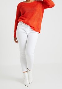 Oranges gestrickter Pullover mit langen Ärmeln, kombiniert mit weißen engen Jeans und weißen Knöchelstiefeln. Die Textur des Pullovers ist grob gestrickt.