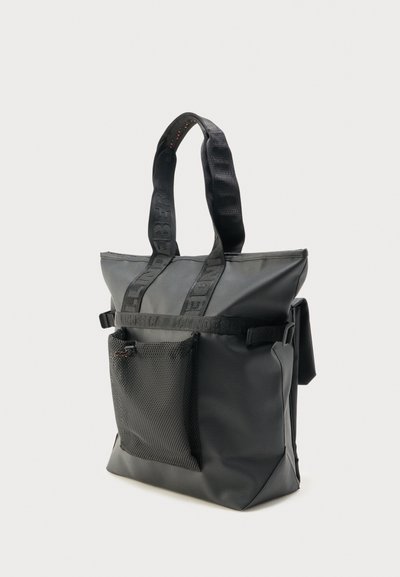 Sac cabas noir avec poche avant en mesh, anses en tissu avec texte, et sangle latérale avec boucle sur un fond clair uni.