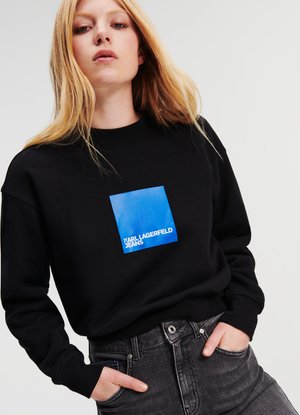 Crna dukserica s plavim kvadratnim grafikama i natpisom "KARL LAGERFELD JEANS." Materijal je mekan, s opuštenim krojem i rebrastim manžetama.