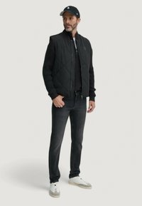 Gilet trapuntato nero sopra un maglione nero, abbinato a jeans scuri e sneakers bianche. L'outfit include un berretto nero con dettagli.