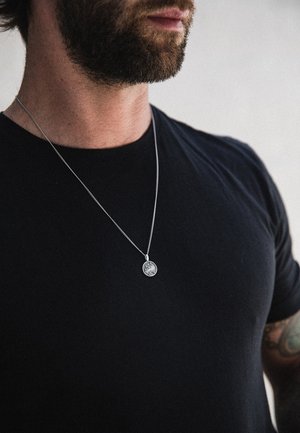 Silver necklace with a round pendant worn over a black t-shirt. Close-up on a male model's neck. The pendant features intricate detailing.