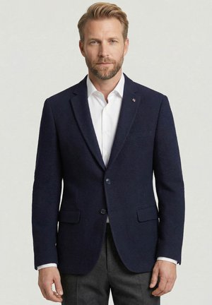 Man met blond haar en baard die een donkerblauw colbert, wit overhemd en donkere broek draagt tegen een effen lichte achtergrond.