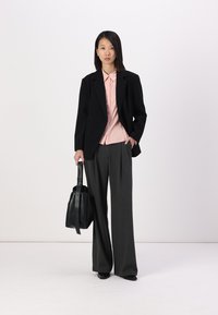 Svart blazer over en lys rosa skjorte med krage, kombinert med grå vide bukser. Holder en svart totebag, satt mot en hvit bakgrunn.