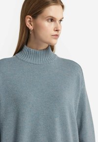 Ljusblå tröja med hög ribbad halsringning, mjuk textur och avslappnad passform. Tyget verkar vara mysigt och lätt med subtil variation.