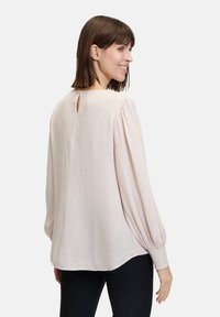 Blusa rosa pallido con lunghe maniche a sbuffo, texture morbida, dettaglio della schiena a chiave e polsini elasticizzati, indossata con pantaloni scuri.