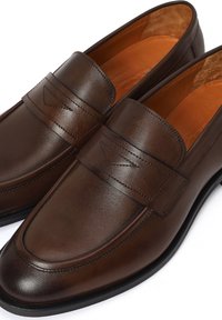 Par de mocasines para hombre de cuero marrón oscuro con suelas negras y costuras decorativas en la correa, vistos desde un ángulo frontal.