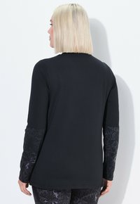 Zwart langemouwtop met gemusterde stof op de mouwen. Heeft een ronde halslijn en een relaxte pasvorm. Materiaal lijkt glad en rekbaar.