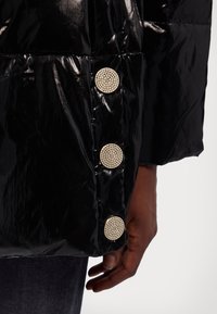 Chaqueta de plumas negra con acabado brillante, botones circulares metálicos con acentos de strass y textura acolchada.