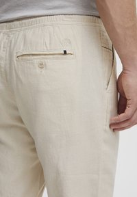 Pantalones de color beige claro hechos de tela texturizada, con un bolsillo trasero con cierre de botón y un pequeño detalle en negro.