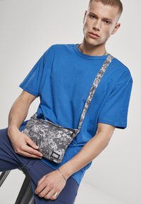 Geantă crossbody gri cu model de camuflaj, cu fermoar negru, purtată peste un tricou albastru, având un material texturat și curea ajustabilă.