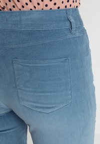 Primer plano de una persona que lleva pantalones de pana azul claro con un bolsillo trasero y una blusa rosa con lunares negros.