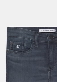 Jeans de denim azul escuro com um pequeno logo bordado "CK" no bolso da frente e uma cintura rotulada. Textura suave e design clássico.