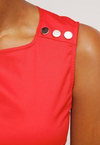 Rote ärmellose Bluse mit rundem Ausschnitt und drei dekorativen Metallknöpfen an der linken Schulter, aus glattem Stoff.