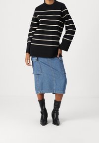 Personne portant un haut à manches longues rayé noir et blanc, une jupe midi en denim bleu avec de grandes poches, et des bottines à talons noires sur fond blanc.